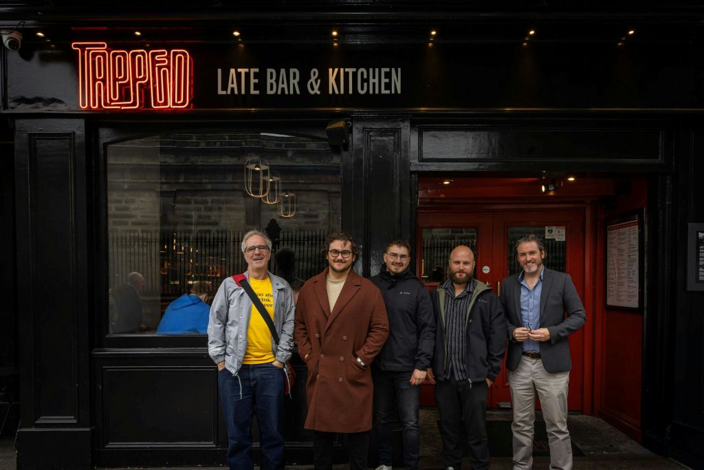 '갇힌 늦은 바 & 키친' 이라는 네온 사인이 있는 검은색 상점 앞에 네 사람이 서 있습니다.