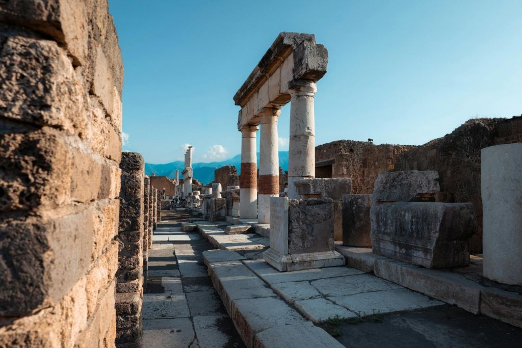 맑고 푸른 하늘 아래 높은 기둥과 부분적인 구조물이 있는 고대 석조 유적지로, 배경에는 산이 보입니다.