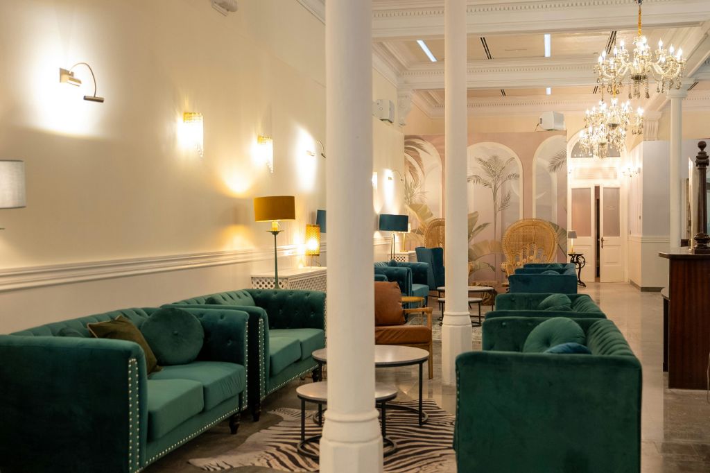 그린 벨벳 소파, 원형 테이블, 벽걸이 램프의 부드러운 조명이 있는 우아한 라운지. 창백한 벽과 기둥이 클래식한 감각을 더합니다.