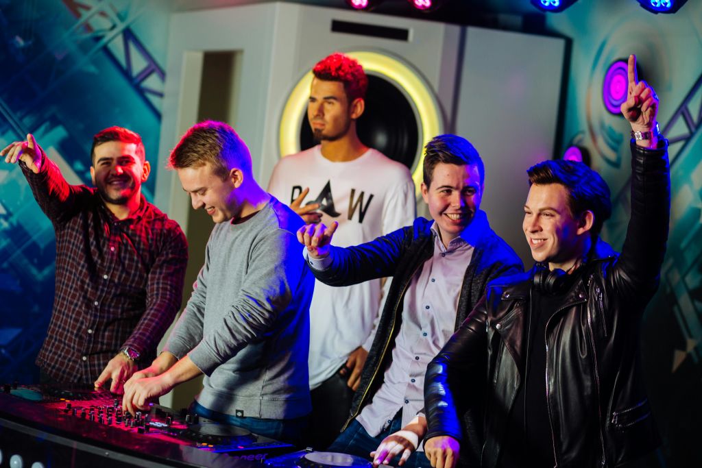 다채로운 색상의 방에 다섯 명의 남성이 있고, 한 명은 디제잉을 하고, 다른 한 명은 웃으며 지켜보고 있습니다.