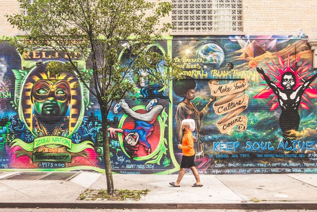 주황색 셔츠를 입은 사람이 다양한 인물, 대담한 텍스트, 생동감 넘치는 우주 예술 작품이 있는 다채로운 벽화를 지나갑니다.