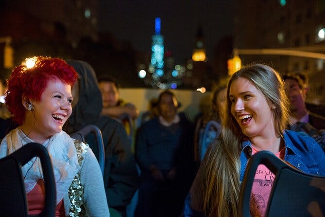 빨간 머리를 한 사람이 다른 사람들과 함께 야간 시티 투어 버스에 앉아 미소를 짓고 있습니다. 배경에는 도시의 불빛과 건물이 있습니다.