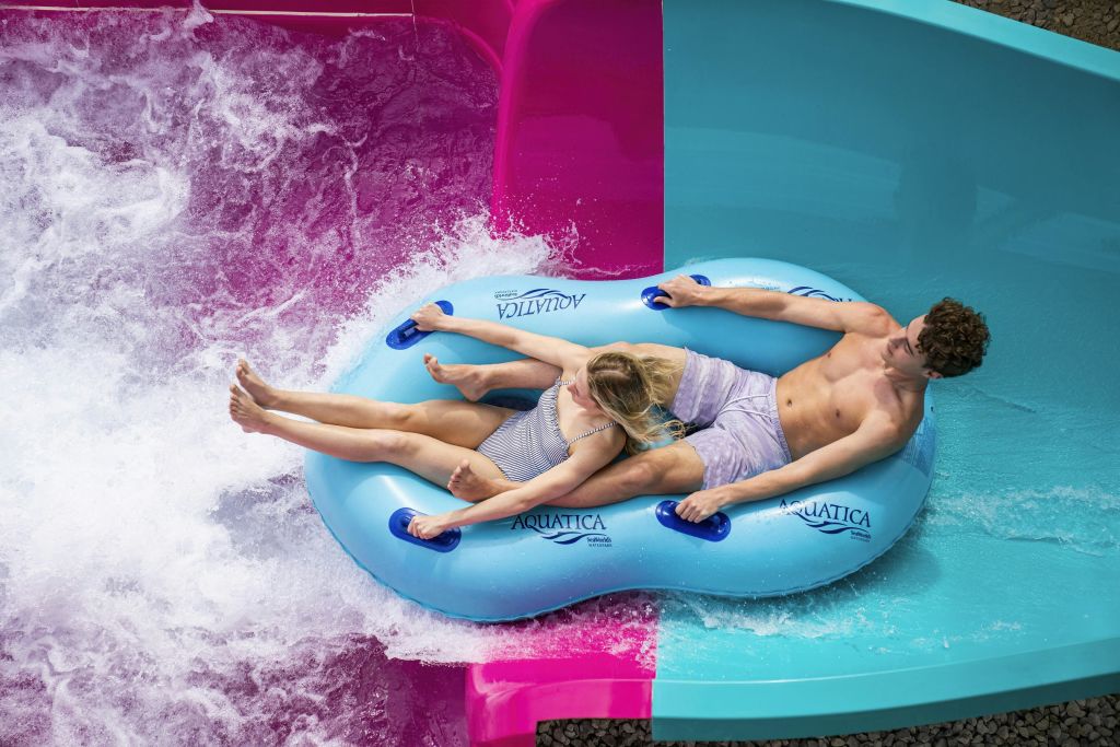 워터파크에서 파란색 팽창식 뗏목을 탄 두 사람이 워터 슬라이드를 따라 분홍색과 파란색으로 나뉘어 미끄러지고 있습니다.