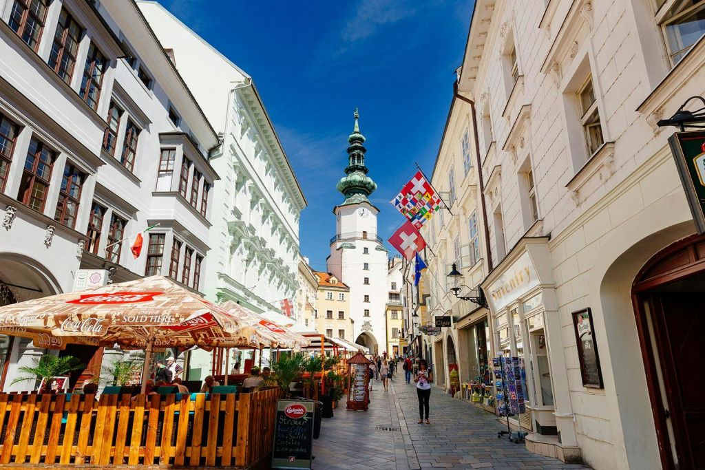 야외 카페, 상점, 산책하는 사람들, 맑고 푸른 하늘 아래 화려한 깃발이 있는 역사적인 타워가 있는 보행자 거리.