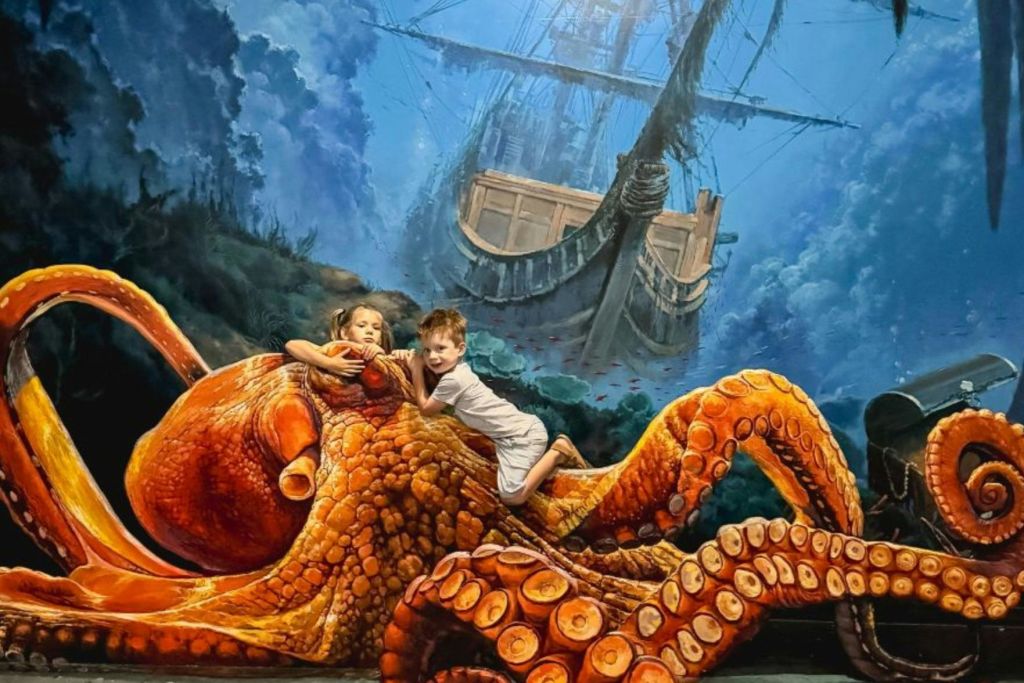 두 어린이는 수중 테마 환경에서 문어와 난파선의 사실적인 3D 아트 디스플레이와 상호 작용합니다.