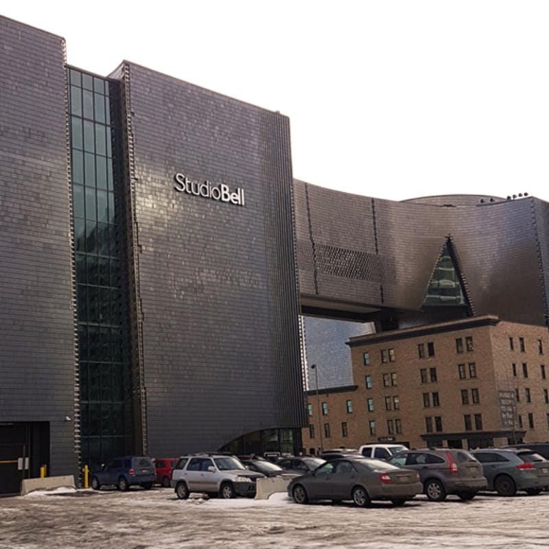 독특한 디자인, 인접한 오래된 구조, 눈으로 뒤덮인 주차 차량이 앞에 있는 "스튜디오 벨" 로 표시된 현대적인 건물.