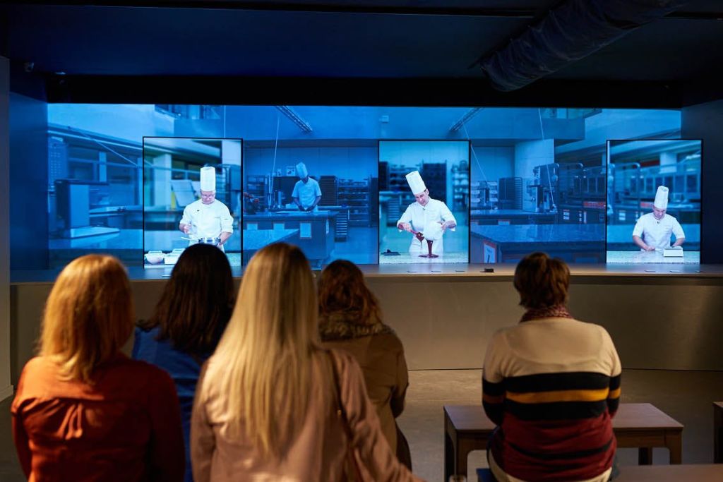 주방에서 셰프의 세 가지 이미지를 보여주는 대형 스크린을 마주하고 앉아 있는 사람들도 있습니다.