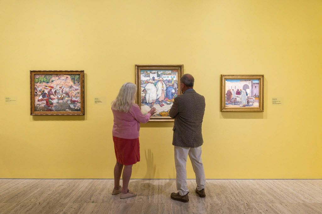두 사람이 갤러리의 노란색 벽에 걸린 액자에 담긴 그림을 관찰합니다.