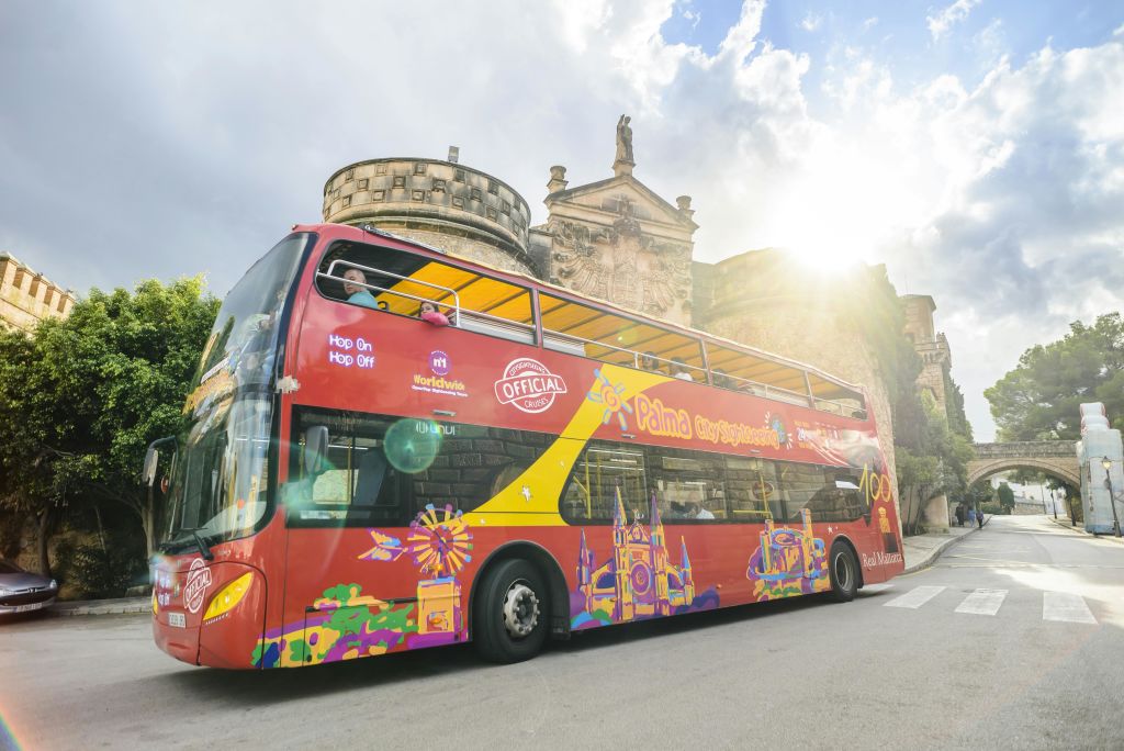 화려한 디자인과 "Hop On Hop Off" 표지판이 있는 밝은 빨간색 2층 관광 버스가 부분적으로 흐린 하늘 아래 역사적인 건물 근처에 주차되어 있습니다.