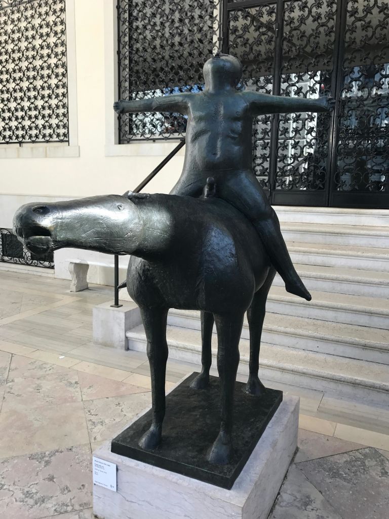 말을 타고 팔을 뻗은 사람의 청동 동상, 계단 근처에 위치, 화려한 건물 외관.