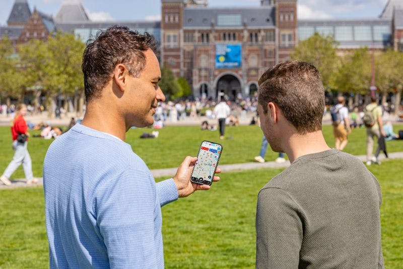 두 남자가 공원에 서 있고, 한 남자는 지도가 표시된 스마트폰을 들고 있습니다. 아치형 통로가 있는 커다란 건물이 배경에 있습니다.