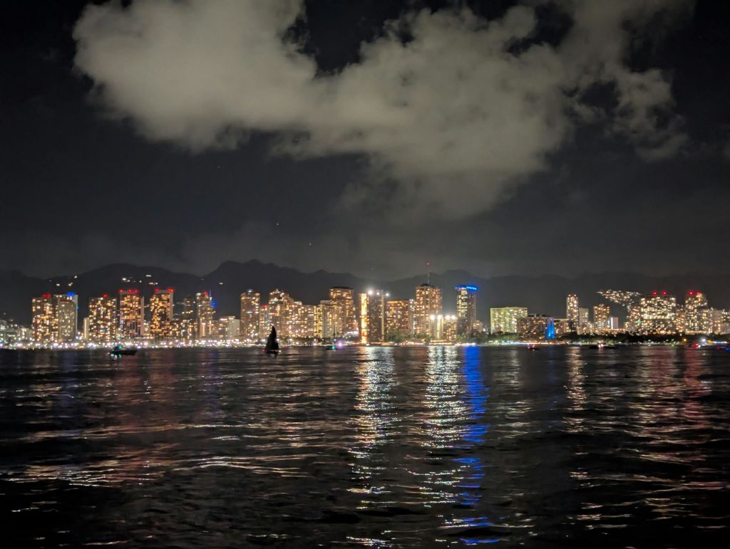 야간에 흐린 하늘 아래 물에 비치는 조명이 있는 건물이 있는 도시의 스카이라인으로, 전면에 실루엣이 있습니다.