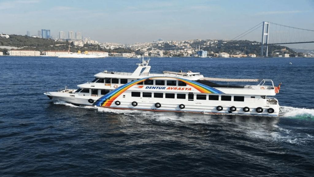 "Dentur Avrasya" 라는 무지개 디자인의 페리가 도시 풍경과 다리를 배경으로 물줄기를 타고 항해합니다.