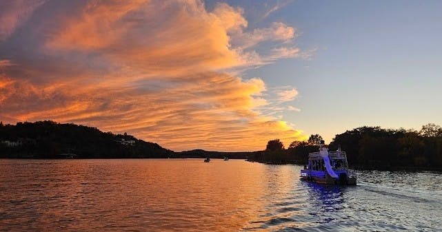 하늘에 극적인 주황색 구름이 드리워진 석양의 고요한 호수는 나무로 둘러싸여 있으며, 오른쪽에는 배가 정박해 있습니다.