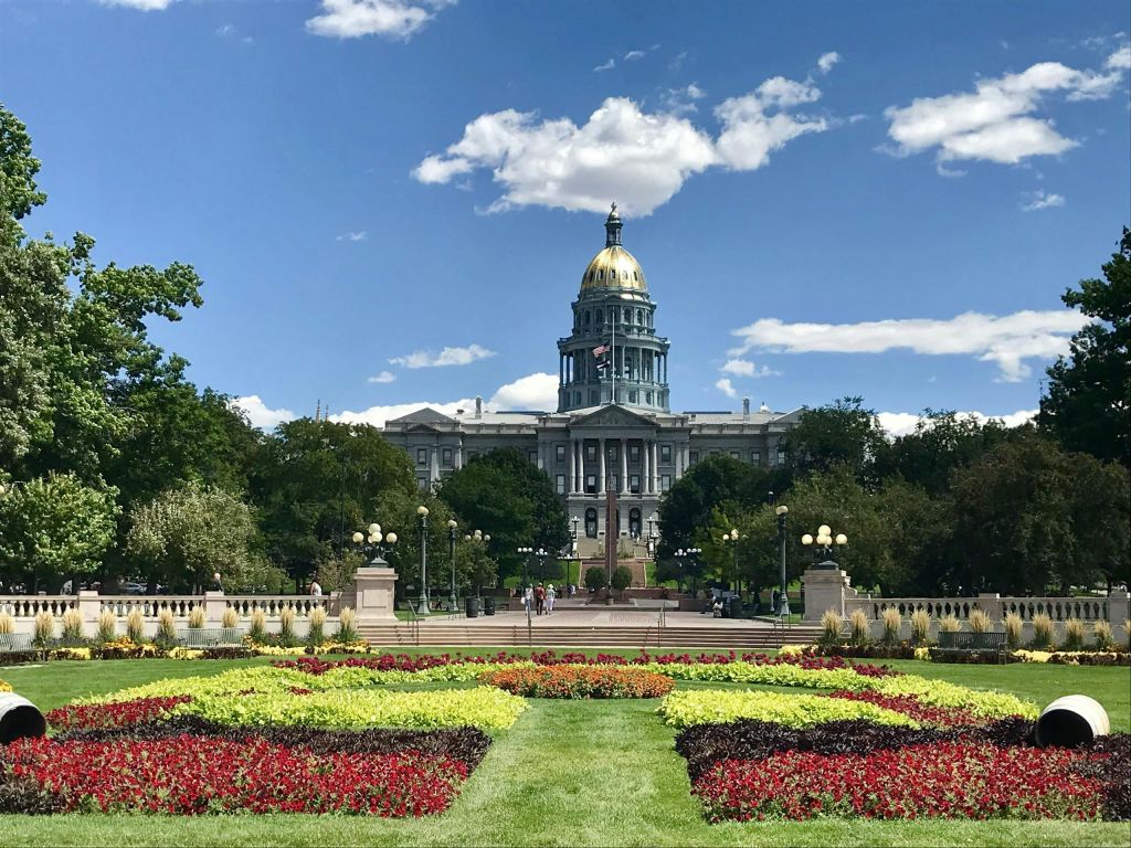 나무로 둘러싸인 황금색 돔이 있는 정부 건물로, 전면에 화려한 꽃 정원이 있습니다.