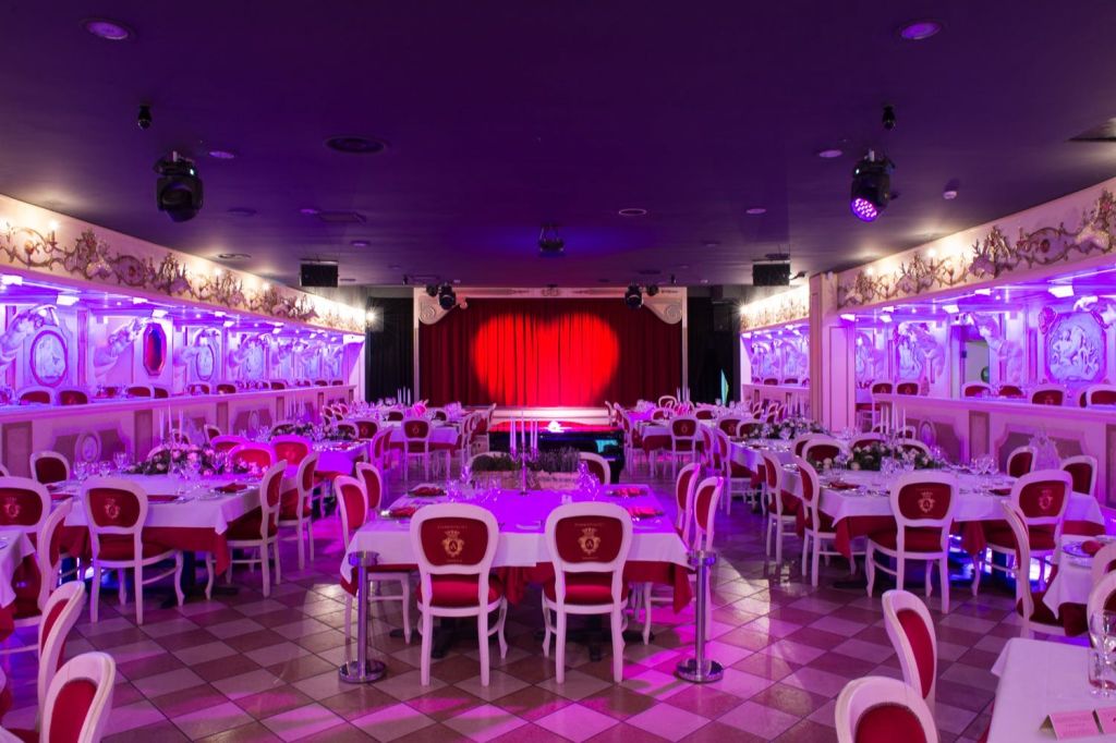 빨간색과 흰색을 테마로 한 인테리어가 돋보이는 연회장, 식사를 위한 원형 테이블, 스포트라이트가 비추는 빨간색 커튼이 있는 무대.