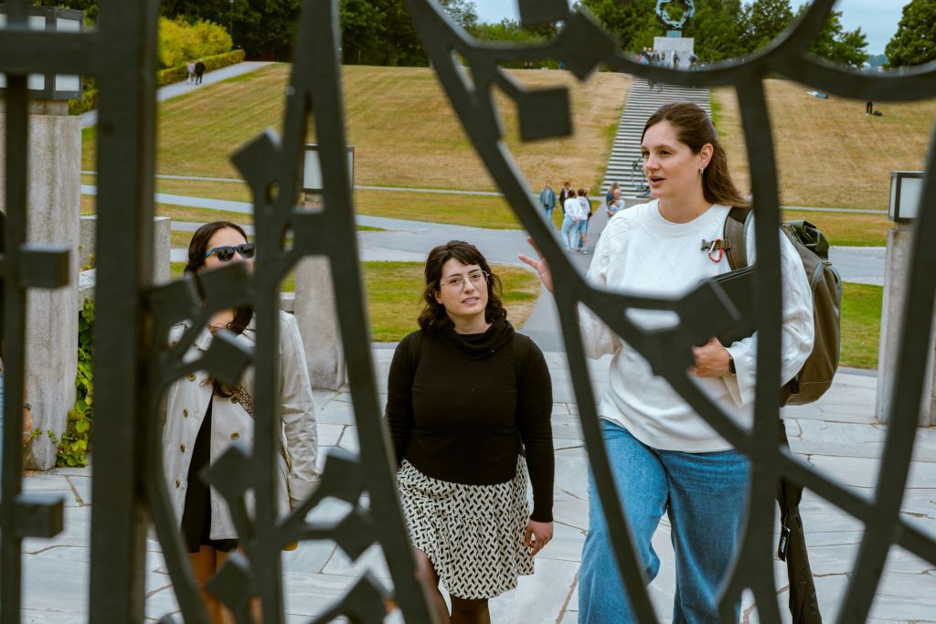 세 명의 여성이 야외에 서서 대화하고 있으며, 장식용 금속 구조물을 통해 볼 수 있습니다. 계단과 잔디 언덕이 배경입니다.