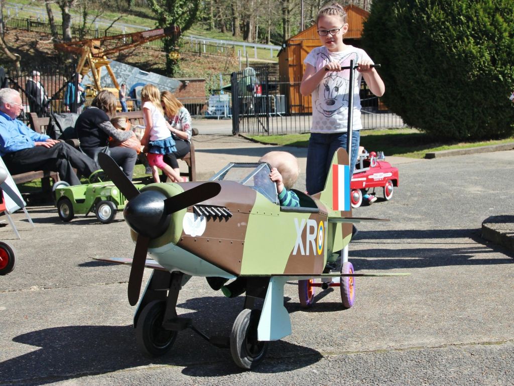 포장된 공간의 작은 장난감 비행기에서 한 소녀가 사람과 장난감 자동차를 배경으로 어린이를 밀고 있습니다.