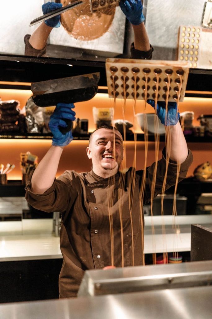 갈색 유니폼을 입은 미소 짓는 셰프가 주황색 조명이 있는 주방에서 파란 장갑을 낀 손으로 캐러멜을 곰팡이에 붓습니다.