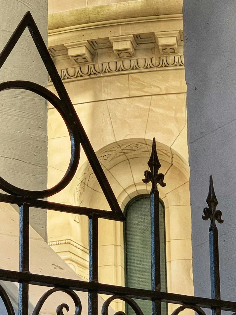 아치형 창문이 있는 유서 깊은 석조 건물 앞에 삼각형과 원형 디자인의 장식용 연철 울타리가 있습니다.