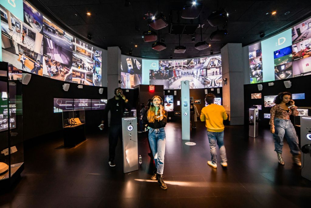 다양한 이미지를 표시하는 여러 개의 대형 화면이 있는 어두운 방에서 사람들은 오디오 방송국과 상호 작용합니다.