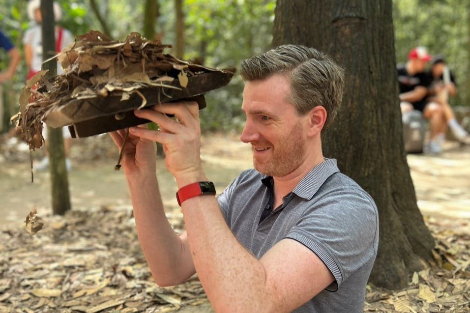 짧은 수염과 회색 셔츠를 입은 남성이 나뭇잎으로 덮개를 들어 올립니다. 그는 야외에 있는 커다란 나무 근처에 서 있습니다.