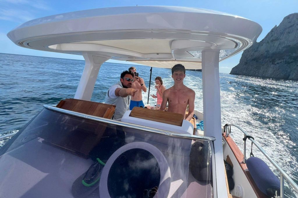 맑은 하늘 아래 보트에 세 사람이 타고 있습니다. 한 사람은 조종하고 다른 두 사람은 사진을 찍고 있습니다. 배경에 바위가 많은 해안선이 보입니다.