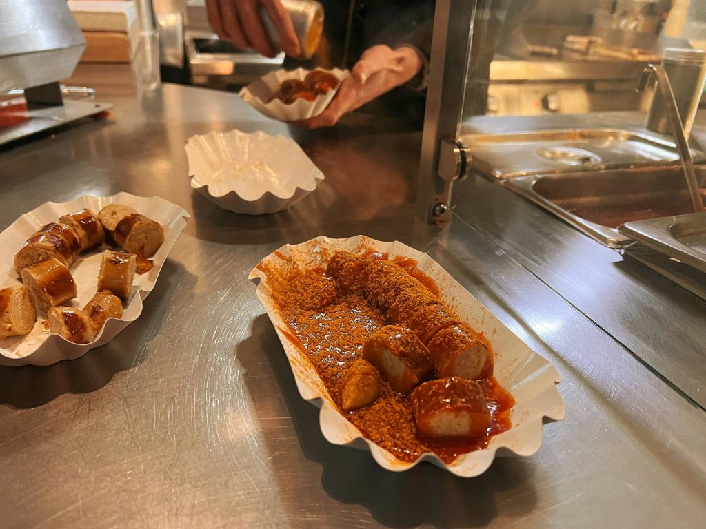 카레 소스와 향신료로 덮인 슬라이스 소시지는 금속 조리대에 담겨 하얀 종이 트레이에 담겨 제공되며, 손으로 양념을 더합니다.