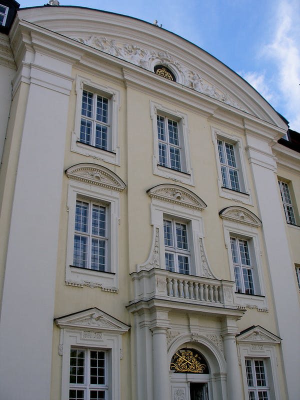 아치형 창문, 화려한 장식, 푸른 하늘을 배경으로 한 작은 발코니가 있는 우아한 베이지색 건물.