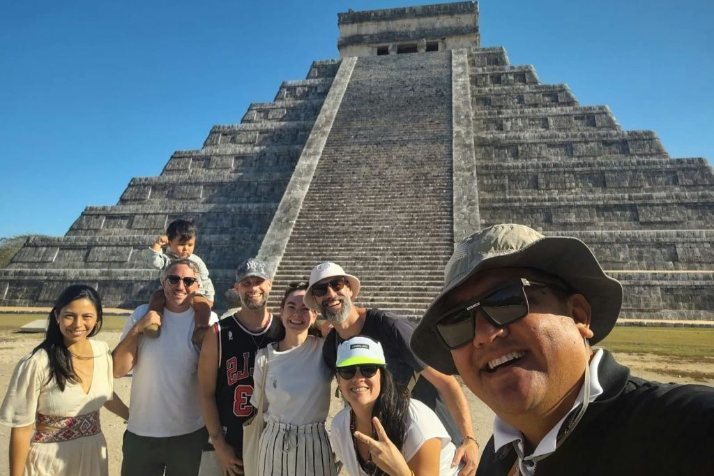 맑고 푸른 하늘 아래 고대의 계단식 피라미드 앞에서 미소를 짓고 포즈를 취하고 있는 한 무리의 사람들.