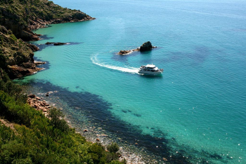 울창한 녹지와 작은 섬이 있는 바위 해안 근처의 맑은 청록색 바다에서 항해하는 보트입니다.