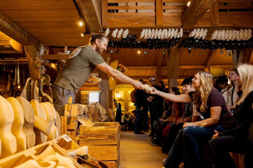 한 남성이 신발이 걸려 있는 작업장에서 앉은 여성에게 나무 신발을 건네고, 사람들이 교환을 지켜보고 있습니다.