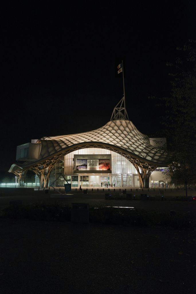 독특하고 물결 모양의 지붕이 있는 현대적인 건물은 밤에 조명이 켜져 있으며, 상단에 깃발이 있고 나무가 부분적으로 오른쪽에 보입니다.