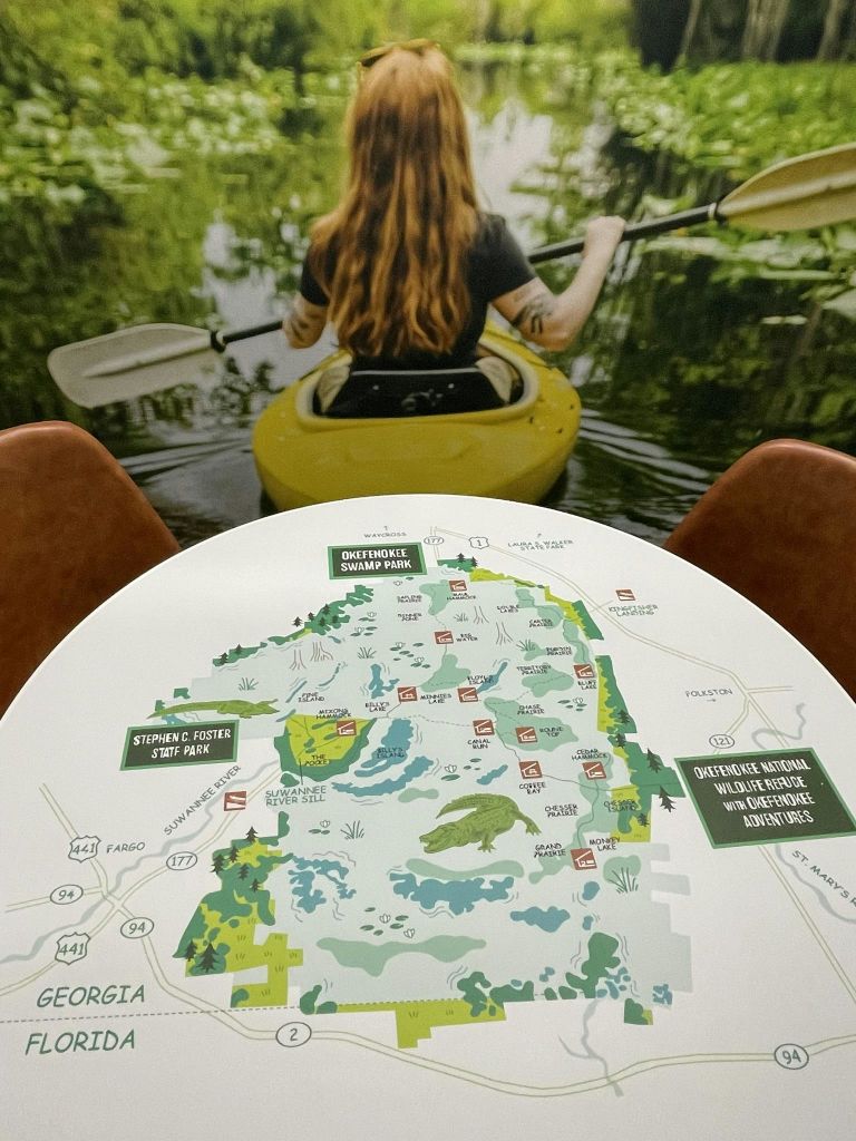 뒤에서 보이는 물 위에서 카약을 타는 여성. 아래는 테이블 위에 오케페노키 늪 공원의 지도이며, 텍스트와 일러스트가 있습니다.