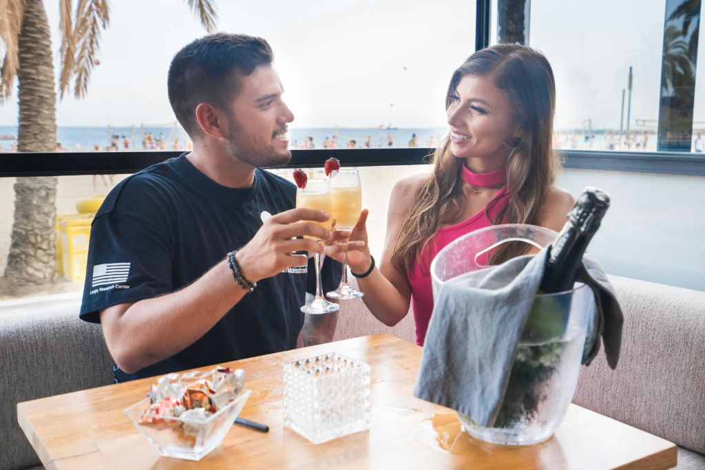 한 남자와 한 여자가 야자수와 바다를 배경으로 해변 테이블에서 칵테일을 마시고 있습니다.