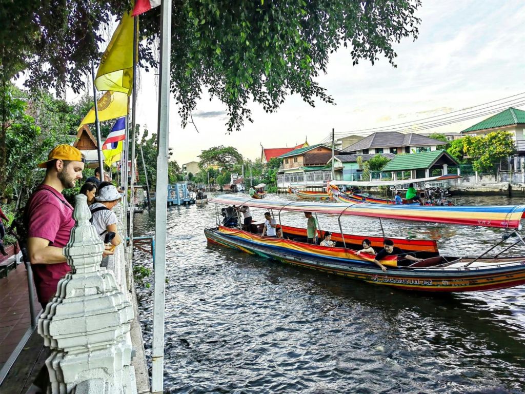 사람들이 강변 산책로를 따라 서서 물 위의 긴 꼬리 보트를 바라보고 있습니다. 산책로에는 깃발과 나무가 늘어서 있습니다.