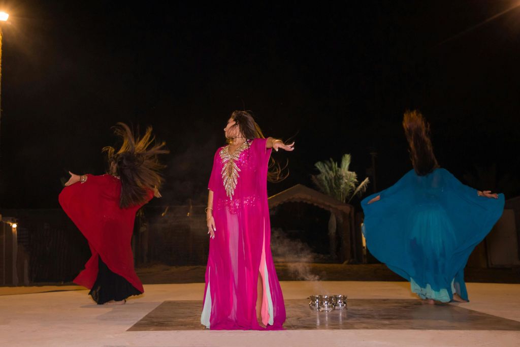 야자수를 배경으로 화려하게 흐르는 의상을 입은 세 사람이 밤에 무대에서 활기차게 춤을 추고 있습니다.