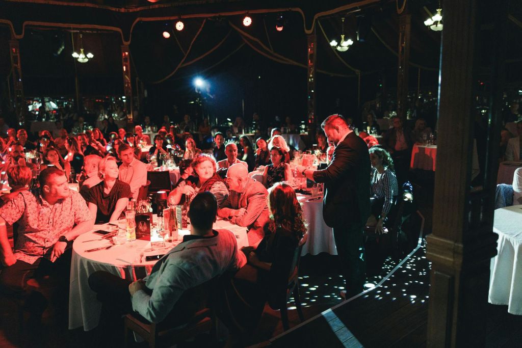 양복을 입은 연사가 빨간색 조명과 스포트라이트로 조명이 희미한 장소의 원형 테이블에 앉아 청중을 응대합니다.