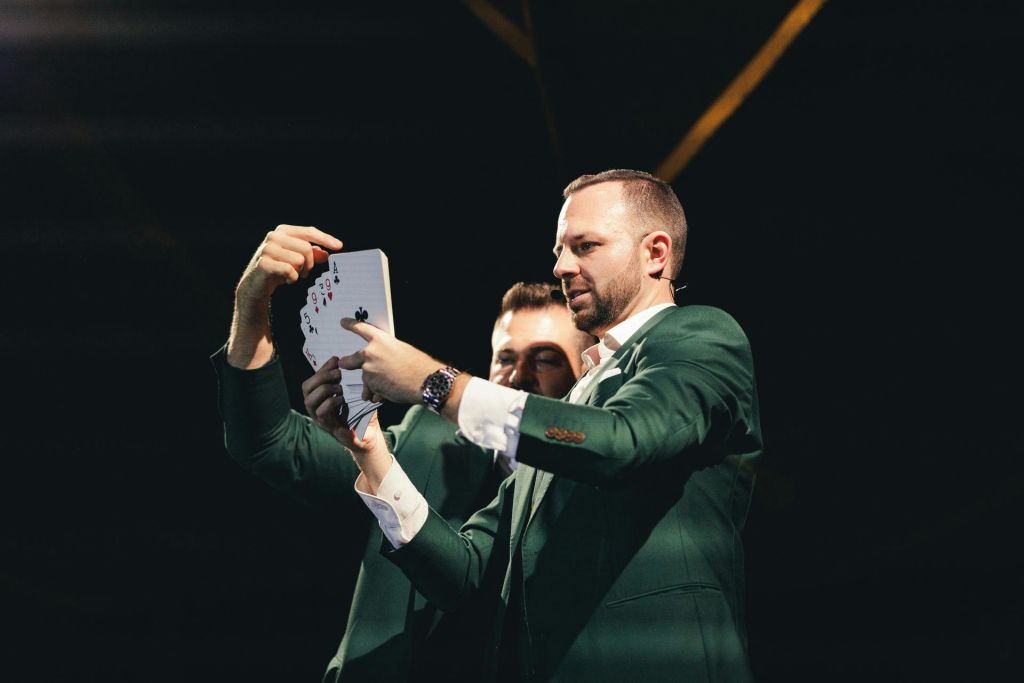 녹색 양복을 입은 두 남자가 무대 조명 아래에서 카드 트릭을 수행하며 카드 놀이의 팬에 중점을 둡니다.