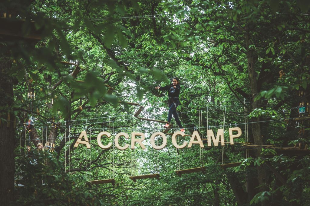 숲이 우거진 어드벤처 파크의 매달린 통나무 다리에서 한 여성이 아래에 커다란 'ACCROCAMP' 문자가 걸려 있는 채 균형을 잡습니다.