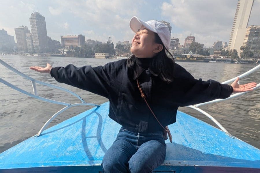 보트에 앉아 양팔을 쭉 뻗은 채 웃고 있는 사람, 도시 풍경과 강을 배경으로 부분적으로 흐린 하늘 아래.