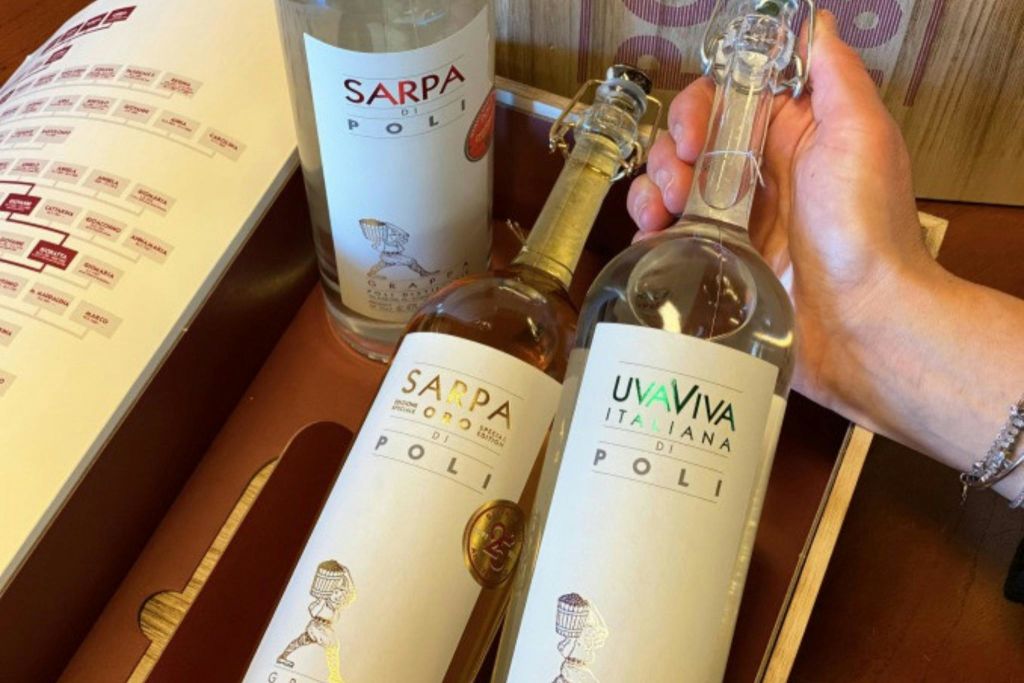 폴리 그라파 3병은 나무 상자에 담겨 있으며, 한 병은 손으로 들고 있습니다. 라벨에는 'Sarpa di Poli', 'Sarpa Oro', 'UvaViva Italiana' 라고 적혀 있습니다.