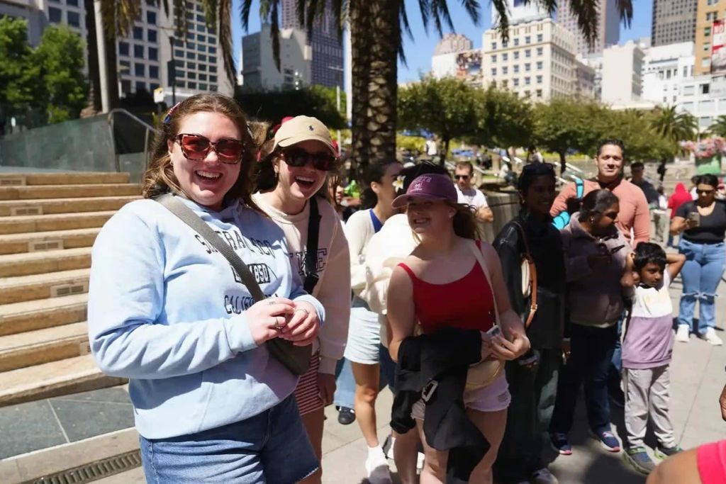 세 사람이 야외에서 카메라를 보며 미소를 짓고 있습니다. 캐주얼한 옷을 입고 야자수와 건물이 있는 화창한 날을 즐기고 있습니다.