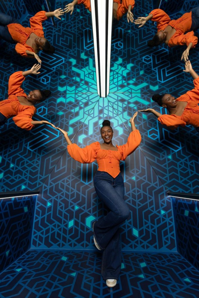 주황색 블라우스와 청바지를 입은 사람이 기하학적 파란색 패턴과 반사가 있는 미러룸에서 포즈를 취하고 있습니다.