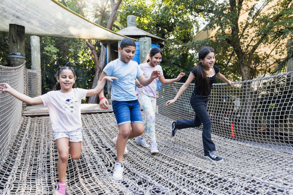 나무와 목조 건물로 둘러싸인 공원의 그물형 다리에서 네 명의 어린이가 웃으며 균형을 잡습니다.