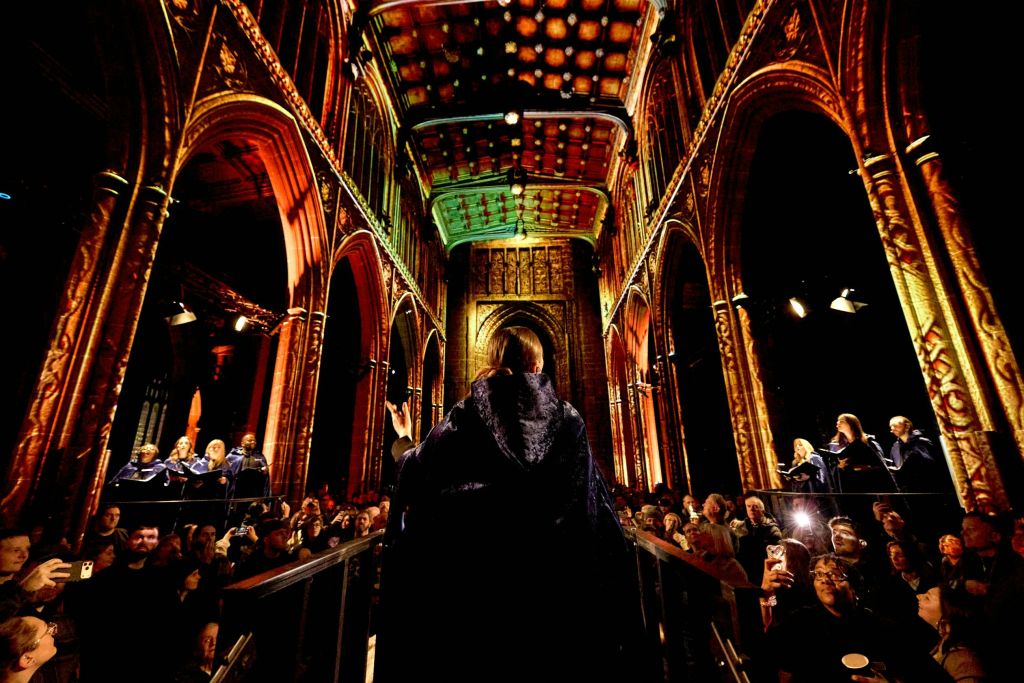 어두운 망토를 입은 사람이 커다랗고 화려한 홀에 아치형 천장이 있는 무대에 서서 관객을 응대하고 있습니다.