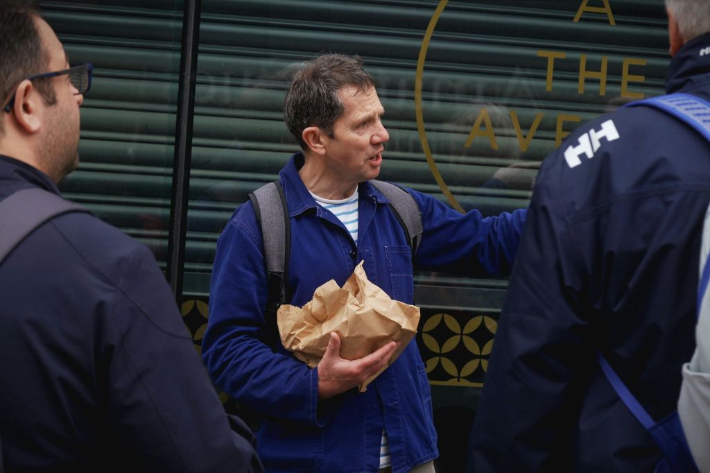 금속 셔터가 닫힌 건물 옆에서 두 남성이 이야기를 나누고 있습니다. 한 남자가 주름진 갈색 종이 봉지를 들고 있습니다.