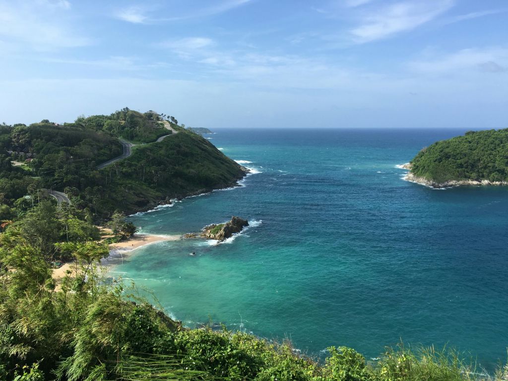 청록색 바다, 바위가 많은 해안, 울창한 푸른 언덕, 푸른 하늘 아래 구불구불한 도로가 펼쳐지는 해안 풍경.