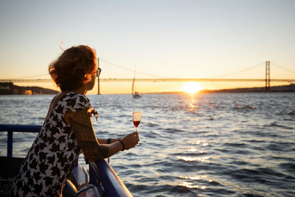 레드 와인 한 잔을 들고 다리를 배경으로 물 위로 지는 석양을 바라보는 문신을 한 사람.