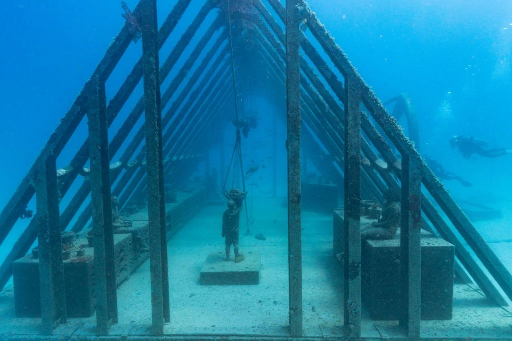 해양 생물로 둘러싸인 금속 프레임워크 아래 인물의 동상이 있는 예배당을 닮은 수중 구조물.